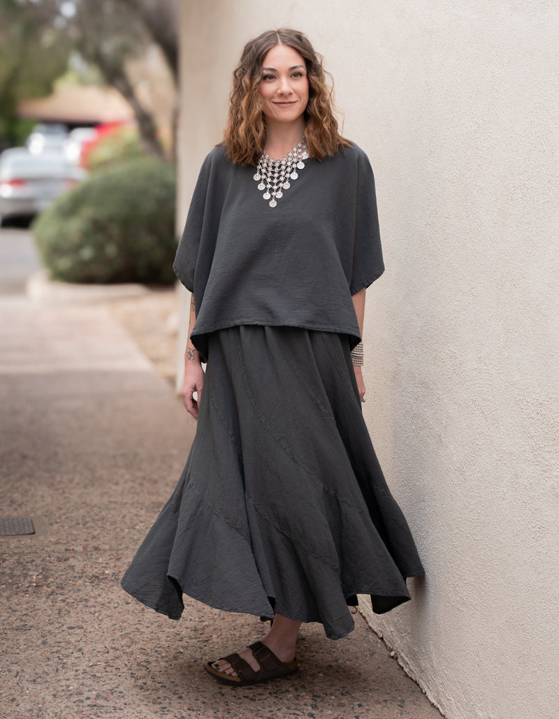 Conscious Coterie woman wearing a dark gray outfit with a necklace, standing against a light-colored wall.