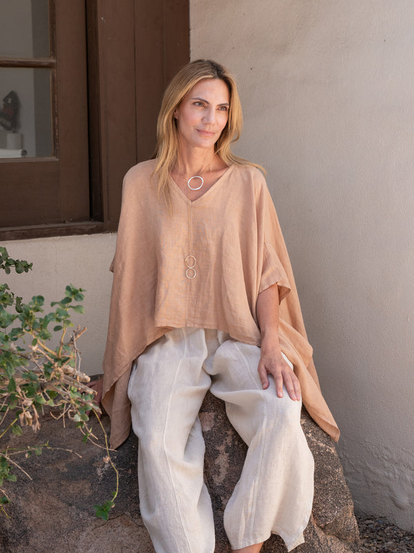 Conscious Coterie Brynn Linen Top in camel on model wearing beige pants sitting against a wall with a plant nearby
