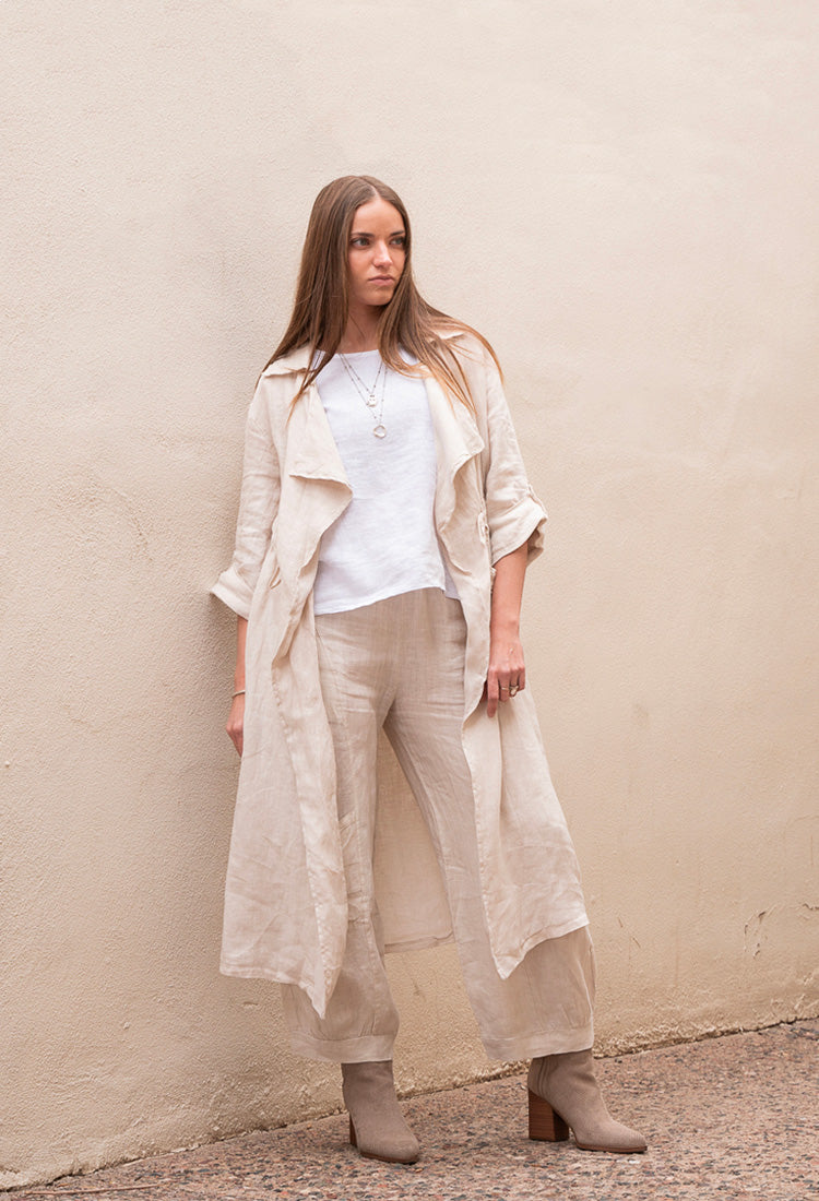 Conscious Coterie womens linen clothing mobile hero image of a woman wearing a long beige coat over a white shirt and pants against a beige wall